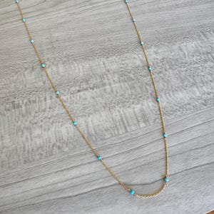 May include: A delicate gold-colored necklace featuring small, evenly spaced turquoise beads. The necklace has a simple chain design and is displayed against a light gray wooden surface. The turquoise beads add a pop of color to the jewelry.
