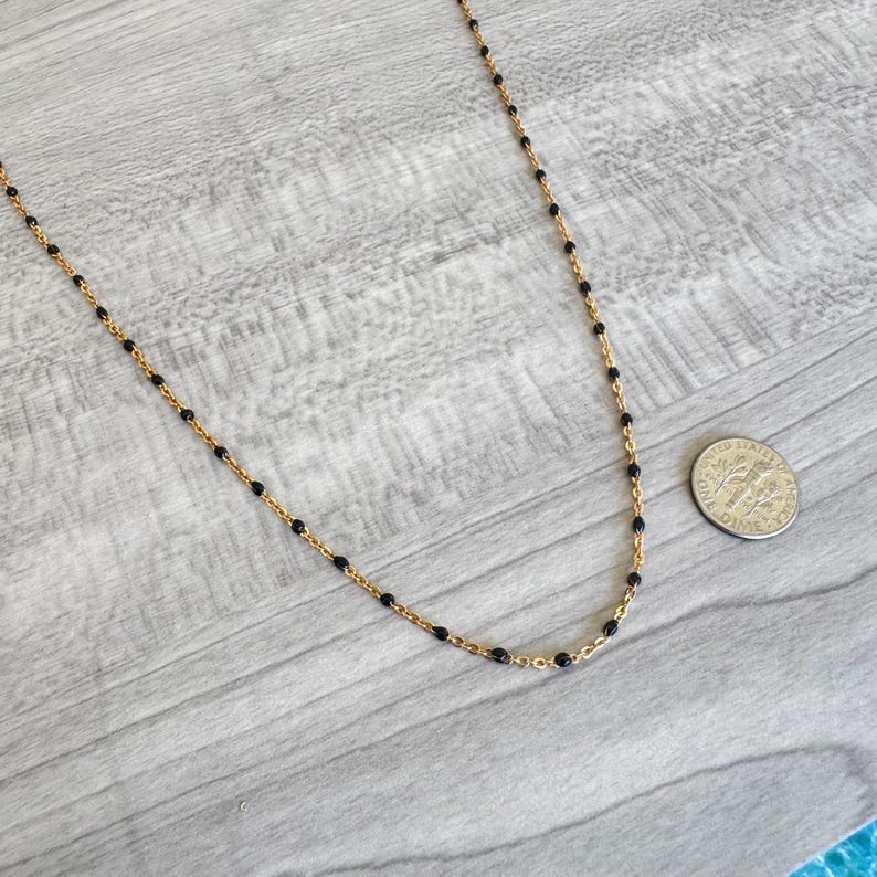 May include: A delicate gold-colored necklace with small black beads evenly spaced along the chain. The necklace is displayed on a light gray wooden surface, with a dime for size reference.