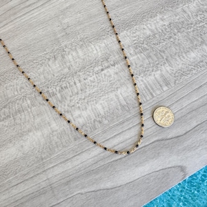 May include: A delicate gold-tone necklace with small black beads evenly spaced along the chain. The necklace is displayed on a light gray wooden surface, with a dime for size reference.