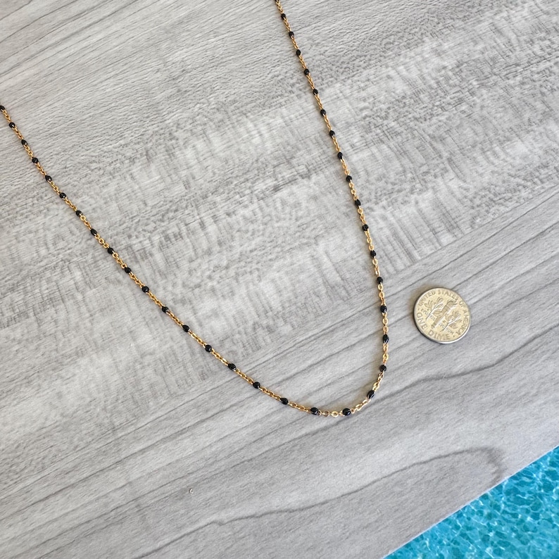 May include: A delicate gold-tone necklace with small black beads evenly spaced along the chain. The necklace is displayed on a light gray wooden surface, with a dime for size reference.