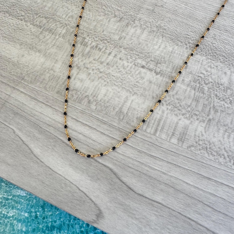May include: A delicate gold-tone necklace featuring small, evenly spaced black beads. The necklace is displayed on a light gray wooden surface, showcasing its simple elegance and design.