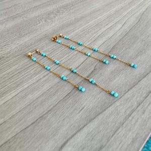 以下が含まれることがあります： ターコイズビーズが付いた長いゴールドのイヤリングのペア。イヤリングは、小さなターコイズビーズがぶら下がった繊細なチェーンで作られています。