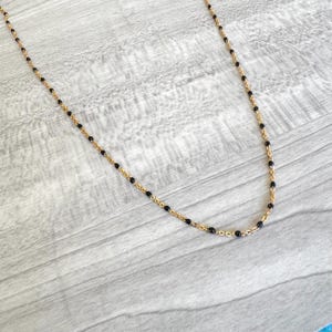 May include: A delicate gold-toned necklace featuring small, evenly spaced black beads. The necklace has a fine chain and a simple, elegant design. The beads are a deep black color, contrasting with the gold chain.