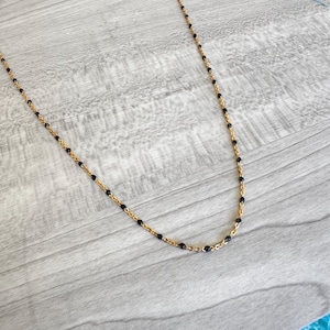 May include: A delicate gold-colored necklace with small, evenly spaced black beads. The necklace is displayed on a light gray wooden surface, showcasing its fine details and elegant design. The chain is thin and the beads are round.