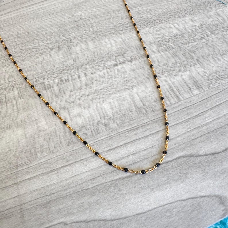 May include: A delicate gold-colored necklace with small, evenly spaced black beads. The necklace is displayed on a light gray wooden surface, showcasing its fine details and elegant design. The chain is thin and the beads are round.