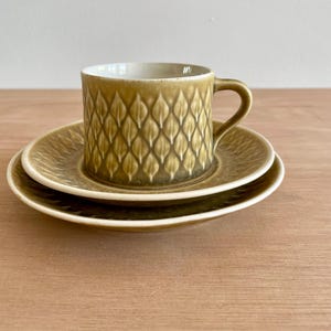 Puede incluir: Un juego de taza y plato de cerámica en tono verde oliva apagado. La taza presenta un patrón repetitivo en relieve de hojas o escamas en el exterior, mientras que el plato muestra una textura de hojas radiantes a juego. El interior es blanco.
