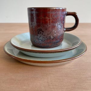 Könnte beinhalten: Ein Keramikbecher mit einer gesprenkelten dunkelroten und braunen Glasur steht auf einem kleinen Teller und einem größeren Teller. Beide Teller haben eine hellgraue Mitte mit einem braunen Rand. Der Becher hat einen zylindrischen Körper.