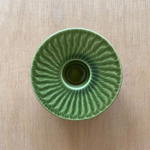 Może przedstawiać: Okrągły zielony świecznik ceramiczny z teksturowanym, promienistym wzorem wypukłych linii biegnących od centralnego otworu do zewnętrznej krawędzi. Szkliwo przechodzi od ciemniejszego odcienia w środku do jaśniejszej zieleni.