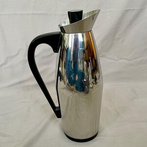 May include: A silver and black stainless steel thermal carafe with a black handle and a lid. The carafe has a shiny, reflective surface.