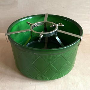 Könnte beinhalten: Ein grüner Weihnachtsbaumständer aus Kunststoff mit einem Metallstützsystem. Der Ständer ist rund mit einem Rautenmuster auf der Außenseite. Die Metallstütze hat verstellbare Arme.