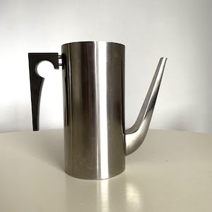 Può includere: Una caffettiera in acciaio inossidabile con un beccuccio lungo e sottile e un manico nero. La caffettiera ha un corpo cilindrico e un design moderno e minimalista. Il manico è attaccato al lato.