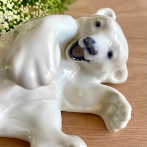 May include: A white porcelain figurine of a polar bear lying on its back with its paws raised. The bear has a glossy finish, dark blue painted eyes, and a dark blue nose and mouth area, capturing a playful pose with its head tilted slightly.