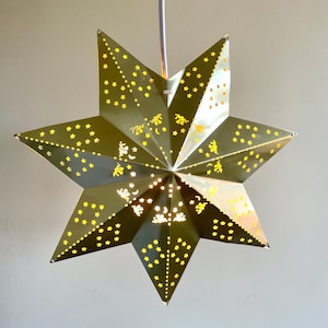 Op de afbeelding: Een zevenpuntige stervormige hanglamp met een metallic gouden afwerking. Het oppervlak is voorzien van ingewikkelde uitsparingen in de vorm van kleine sterren, maansikkels en cherubijntjes, waardoor het licht van binnenuit naar buiten schijnt.
