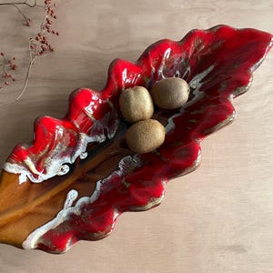 Könnte beinhalten: Eine blattförmige Servierschale aus Keramik mit glänzender roter und brauner Glasur. Die Schale enthält drei Kiwis. Der Rand hat ein wellenförmiges Design mit weißen Akzenten. Ein Zweig mit Beeren befindet sich im Hintergrund.
