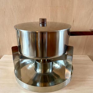 Può includere: Una pentola per fonduta in acciaio inossidabile con manico e pomello del coperchio in legno. La pentola poggia su un supporto abbinato con base circolare. Il design è vintage e adatto all'intrattenimento.