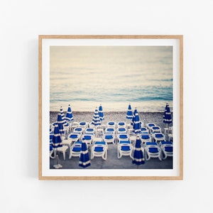 May include: A photograph of a beach scene with rows of blue and white striped beach chairs and umbrellas. The chairs are lined up in rows on the sand, facing the ocean. The water is a light blue color and the sky is a pale blue.