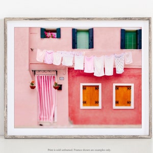 Peut inclure: Un b&acirc;timent rose avec un auvent ray&eacute; au-dessus de la porte et du linge suspendu &agrave; une corde &agrave; linge. Les fen&ecirc;tres sont vertes et les volets sont bleus. Il y a deux plantes en pot accroch&eacute;es au mur.