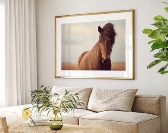 May include: A framed photograph of a brown horse with a flowing mane, standing in a field. The photo is hanging on a white wall in a living room setting with a beige sofa and a potted plant.
