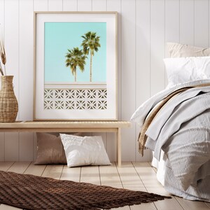 May include: A framed print of two palm trees against a light blue sky. The print is hanging on a white wall above a wooden bench. The bench has two pillows on it. A brown rug is on the floor in front of the bench. A bed with white linens and a brown throw blanket is in the background.