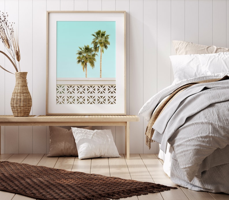 May include: A framed print of two palm trees against a light blue sky. The print is hanging on a white wall above a wooden bench. The bench has two pillows on it. A brown rug is on the floor in front of the bench. A bed with white linens and a brown throw blanket is in the background.