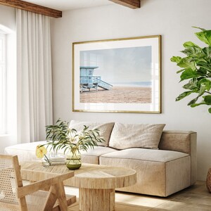 Peut inclure: Un int&eacute;rieur de salon avec un canap&eacute; en L beige clair, une table basse en bois, un fauteuil en osier et une grande photographie encadr&eacute;e d'une cabine de plage sur une plage.