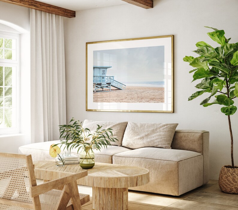 Peut inclure: Un int&eacute;rieur de salon avec un canap&eacute; en L beige clair, une table basse en bois, un fauteuil en osier et une grande photographie encadr&eacute;e d'une cabine de plage sur une plage.