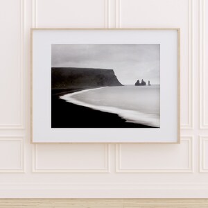 May include: A black and white photograph of a rocky coastline with a dark, sandy beach and a body of water. The sky is overcast and the water is calm.
