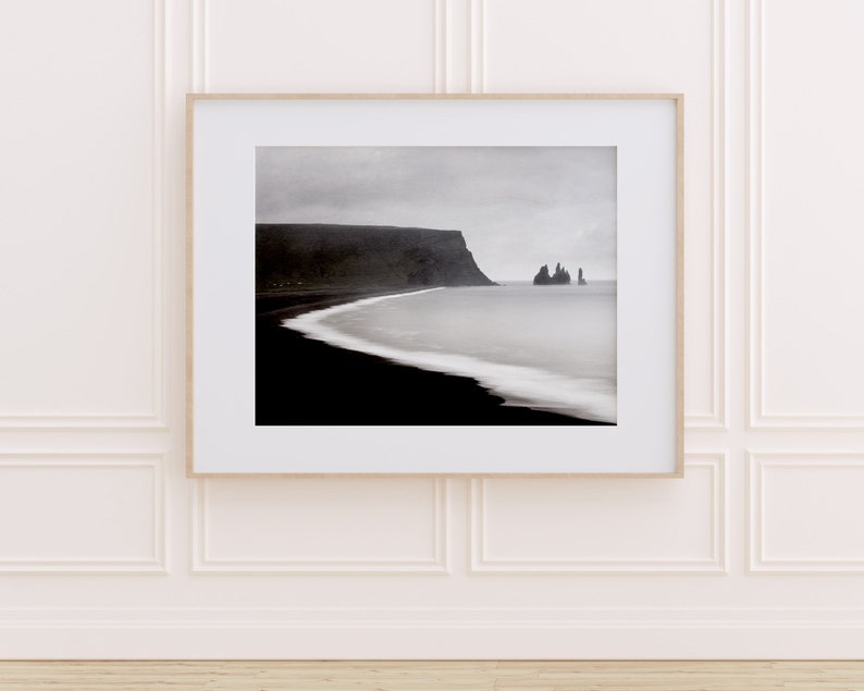 May include: A black and white photograph of a rocky coastline with a dark, sandy beach and a body of water. The sky is overcast and the water is calm.