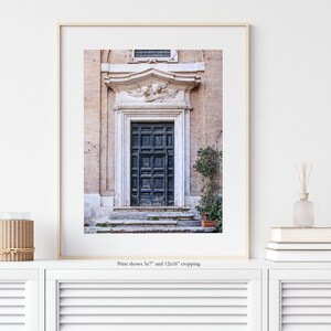 May include: A vintage-style photograph of a dark blue wooden door with a decorative stone archway and a small bush in front. The door is set against a backdrop of a light beige building.