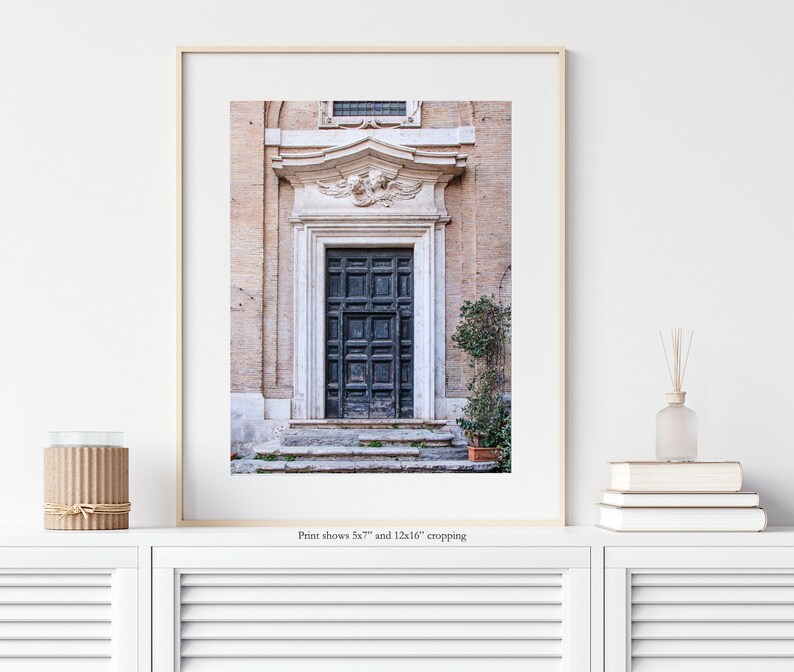 May include: A vintage-style photograph of a dark blue wooden door with a decorative stone archway and a small bush in front. The door is set against a backdrop of a light beige building.