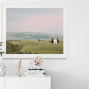 May include: A framed photograph of a white chapel with a pink sky in the background. The chapel is located on a grassy hill with rolling hills in the distance. The photo is framed in a white frame.