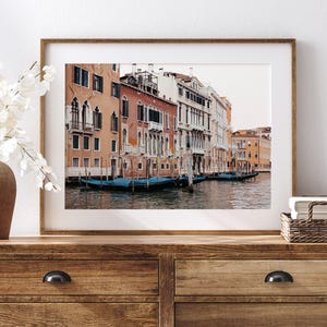 May include: A framed photograph of a canal in Venice, Italy. The photo shows colorful buildings lining the canal, with gondolas docked along the edge. The water is calm and reflects the buildings and sky.