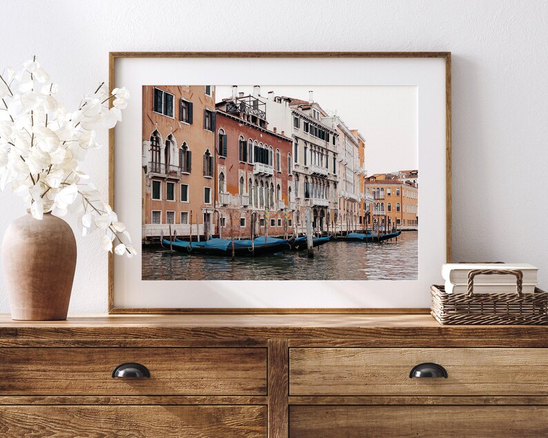 May include: A framed photograph of a canal in Venice, Italy. The photo shows colorful buildings lining the canal, with gondolas docked along the edge. The water is calm and reflects the buildings and sky.
