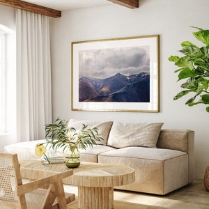 Puede incluir: Un interior de sala de estar con un sof&aacute; beige, una mesa de caf&eacute; de madera, una silla de mimbre y una fotograf&iacute;a enmarcada de una cordillera con un cielo nublado. Una planta en maceta con grandes hojas verdes est&aacute; en la esquina de la habitaci&oacute;n.
