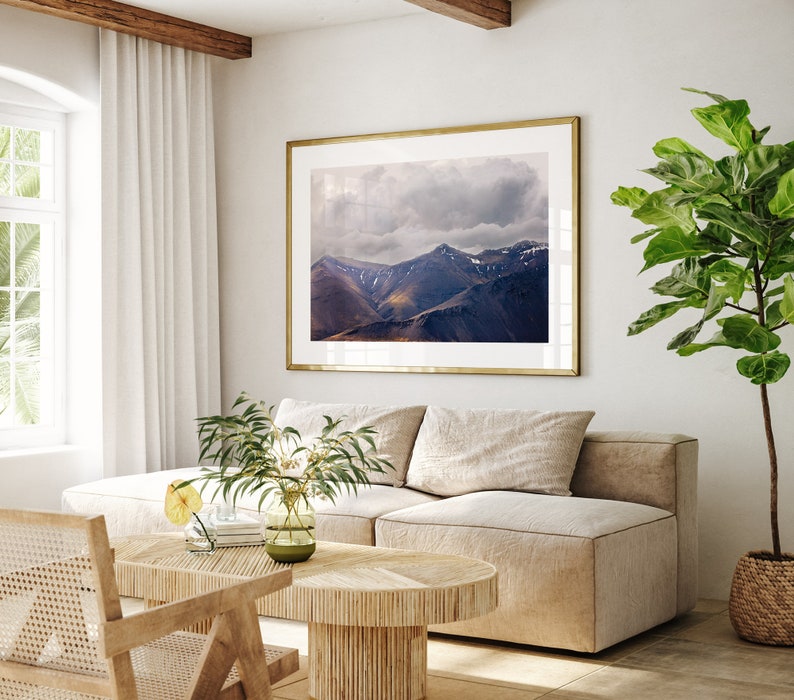 Puede incluir: Un interior de sala de estar con un sof&aacute; beige, una mesa de caf&eacute; de madera, una silla de mimbre y una fotograf&iacute;a enmarcada de una cordillera con un cielo nublado. Una planta en maceta con grandes hojas verdes est&aacute; en la esquina de la habitaci&oacute;n.