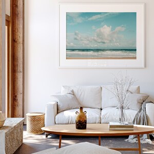 May include: A white couch with two pillows and a throw blanket sits in a living room with a wooden coffee table and a framed photograph of a beach scene with blue sky and white clouds. A brown ceramic vase with a textured surface sits on the coffee table.