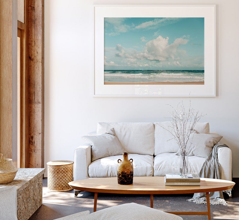 May include: A white couch with two pillows and a throw blanket sits in a living room with a wooden coffee table and a framed photograph of a beach scene with blue sky and white clouds. A brown ceramic vase with a textured surface sits on the coffee table.