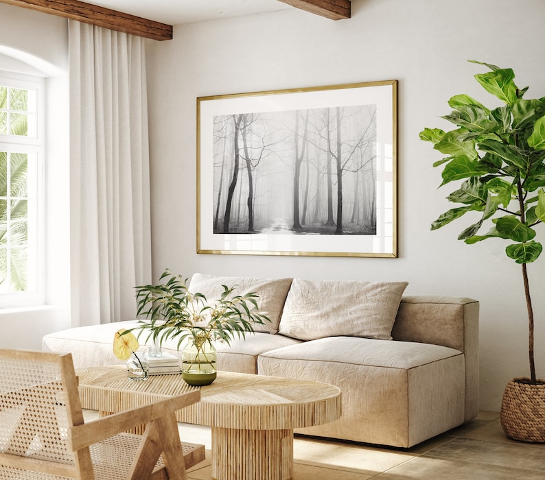 May include: A living room with a beige sectional sofa, a wooden coffee table, a wicker chair, and a large framed black and white photograph of a foggy forest. A potted green plant is in the corner of the room.