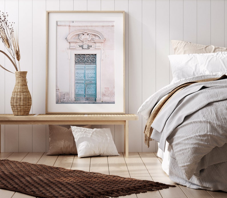 May include: A framed photograph of a vintage blue door with ornate ironwork, set against a pale pink wall. The door is framed by a white archway and is surrounded by a light pink building.