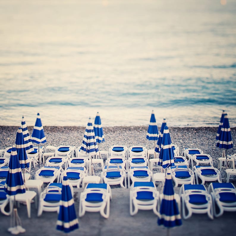 May include: A beach scene with rows of blue and white striped beach chairs and umbrellas lined up on the sand. The chairs are empty and the ocean is in the background.