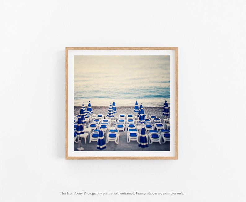 May include: A photograph of a beach scene with rows of blue and white striped beach chairs and umbrellas. The chairs are lined up in rows on the sand, facing the ocean. The water is a light blue color and the sky is a pale blue.