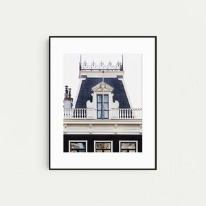 May include: A black and white photograph of a building facade in Amsterdam, Netherlands. The building has a traditional Dutch gable roof with a dormer window and a decorative railing. The building is painted white with black trim.