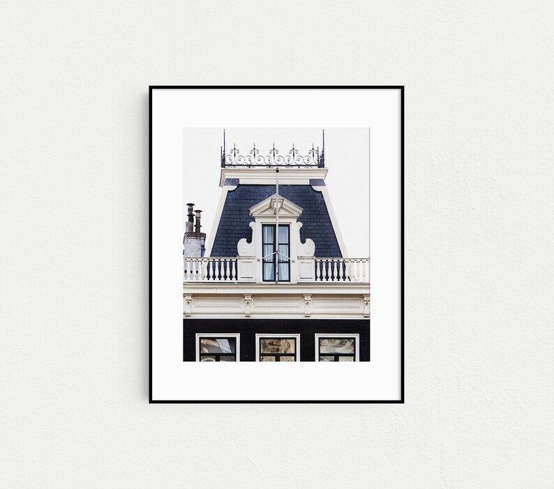 May include: A black and white photograph of a building facade in Amsterdam, Netherlands. The building has a traditional Dutch gable roof with a dormer window and a decorative railing. The building is painted white with black trim.