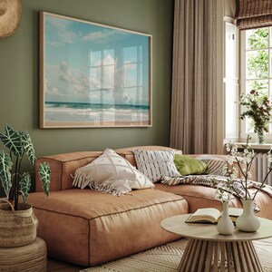 May include: A light brown leather sectional sofa with white and black patterned pillows, a green pillow, and a throw blanket. The sofa is in a living room with a framed beach scene on the wall, a window with white blinds, and a wooden coffee table with a book and two white vases.