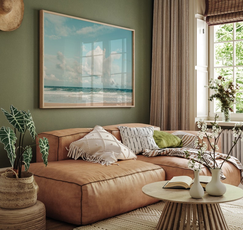 May include: A light brown leather sectional sofa with white and black patterned pillows, a green pillow, and a throw blanket. The sofa is in a living room with a framed beach scene on the wall, a window with white blinds, and a wooden coffee table with a book and two white vases.