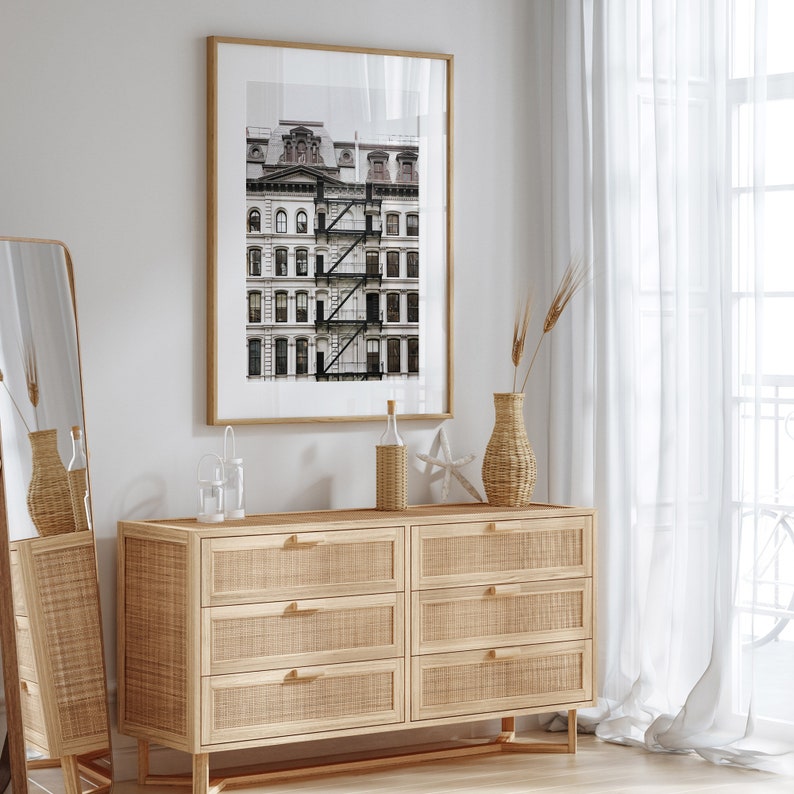 Puede incluir: Una c&oacute;moda de madera clara con seis cajones, un frente de rat&aacute;n tejido y tiradores dorados. La c&oacute;moda est&aacute; en una habitaci&oacute;n con una pared blanca, una fotograf&iacute;a en blanco y negro enmarcada de un edificio y una cortina blanca transparente.