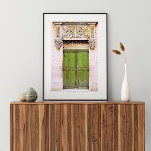 May include: A framed photograph of a green wooden door with ornate carvings above it. The door is set in a pink wall with white trim.
