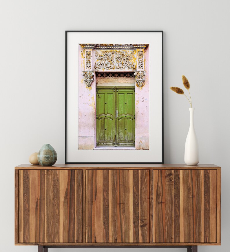 May include: A framed photograph of a green wooden door with ornate carvings above it. The door is set in a pink wall with white trim.
