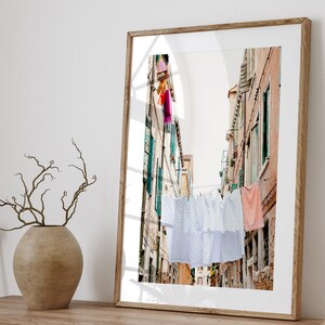 Pu&ograve; includere: Una fotografia incorniciata di un vicolo stretto a Venezia, Italia, con biancheria bianca appesa a uno stendino teso tra gli edifici. Gli edifici sono dipinti in tonalit&agrave; di rosa e giallo, e la biancheria ondeggia dolcemente nella brezza.