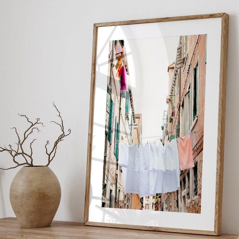 Pu&ograve; includere: Una fotografia incorniciata di un vicolo stretto a Venezia, Italia, con biancheria bianca appesa a uno stendino teso tra gli edifici. Gli edifici sono dipinti in tonalit&agrave; di rosa e giallo, e la biancheria ondeggia dolcemente nella brezza.